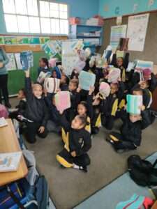 Learners sit on ground, eagerly answer the teacher and holding papers in the air at Primary School in Langebaan