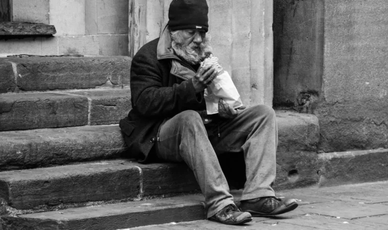 Iihomeless man sits on steps drinking liquor out of a brown bag.