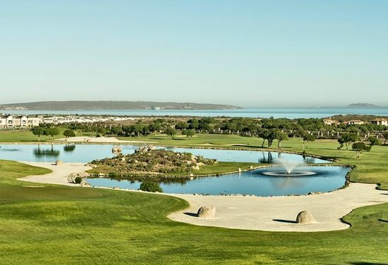 Love weskus ilove weskus igolf course with green fairways, a water pond and fountain, sandy bunkers, and houses in the distance under a clear blue sky.