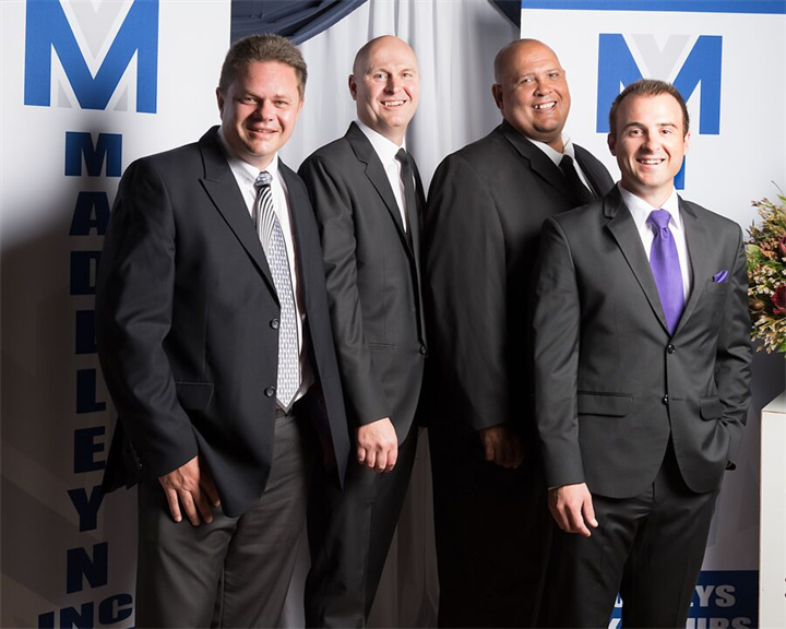 Four men in dark suits posing for a group photo at a corporate event, standing in front of a blue-and-white backdrop with large M lettering.