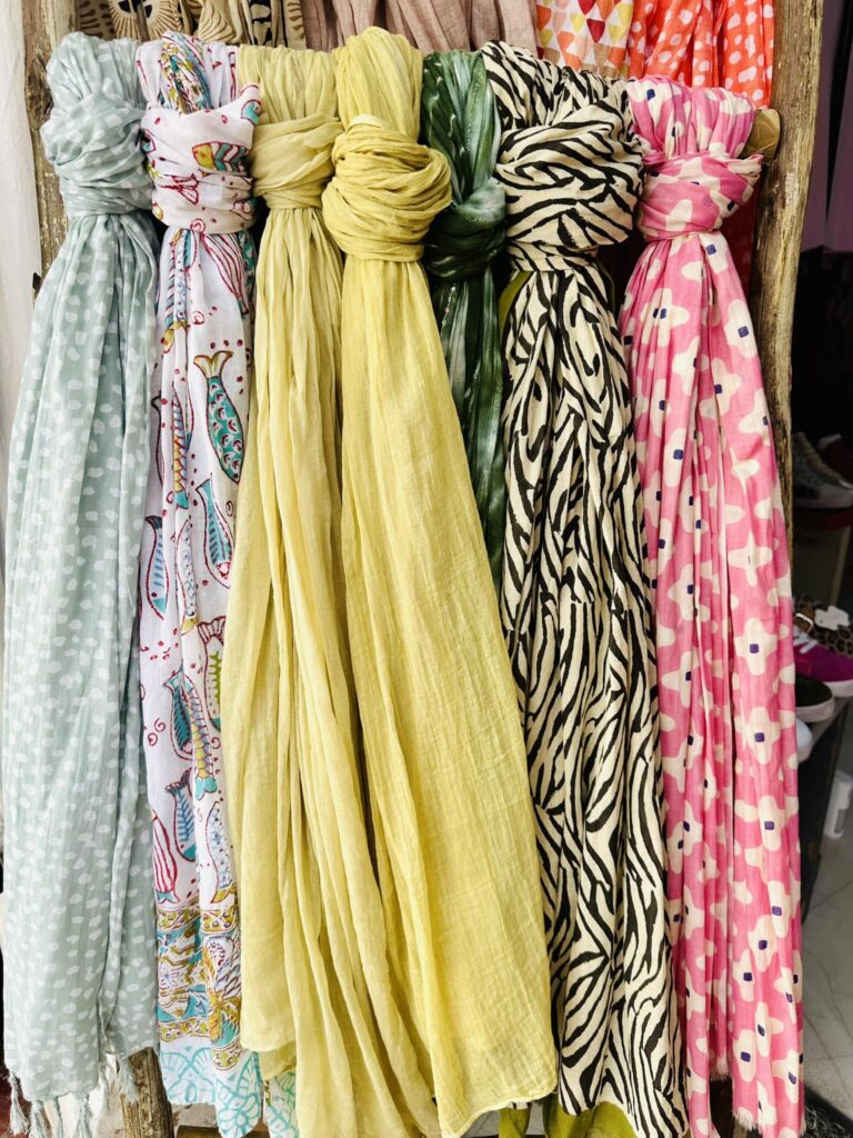 Row of colorful scarves hanging side by side in pastel yellows, greens, zebras, and pink patterns on a wooden rack, displaying fabric patterns and textures. All available at Blooming Nice Boutique in Langebaan.
