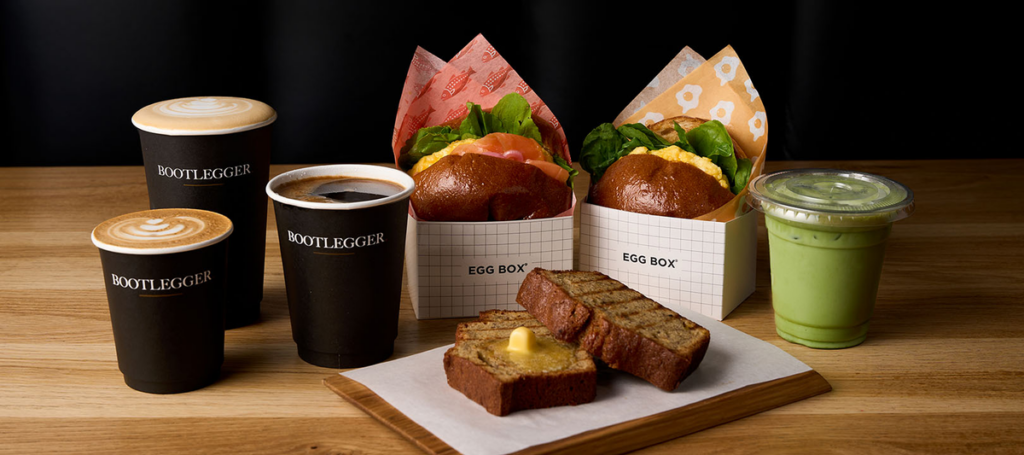 Breakfast set with two egg-box sandwiches, three Bootlegger coffee cups, a green smoothie, and buttered toast on a wooden table.