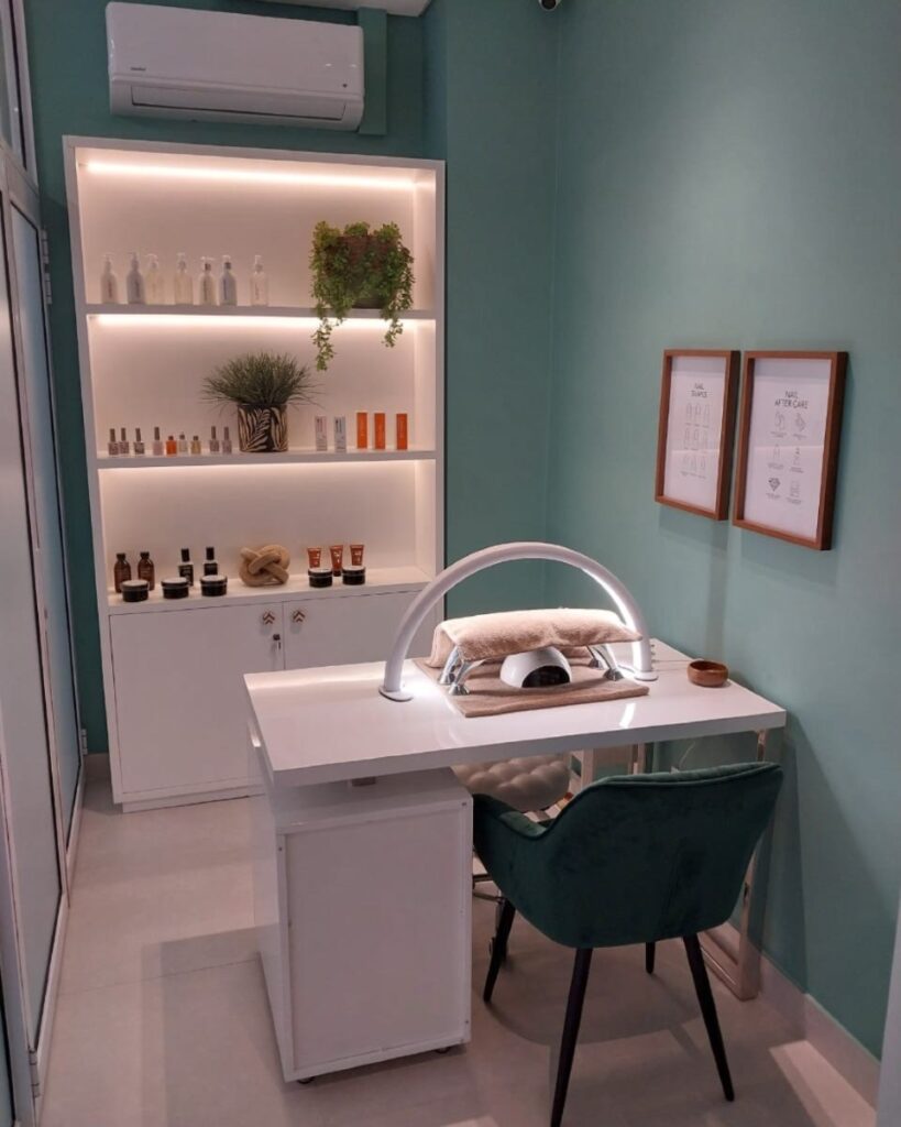Manicure station in a teal-walled spa: white table, curved lamp, green upholstered chair, and shelving with nail products and plants.