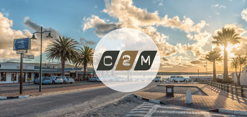 Coastal street at sunset with palm trees, parked cars, and a large circular C2M logo overlay in the center.