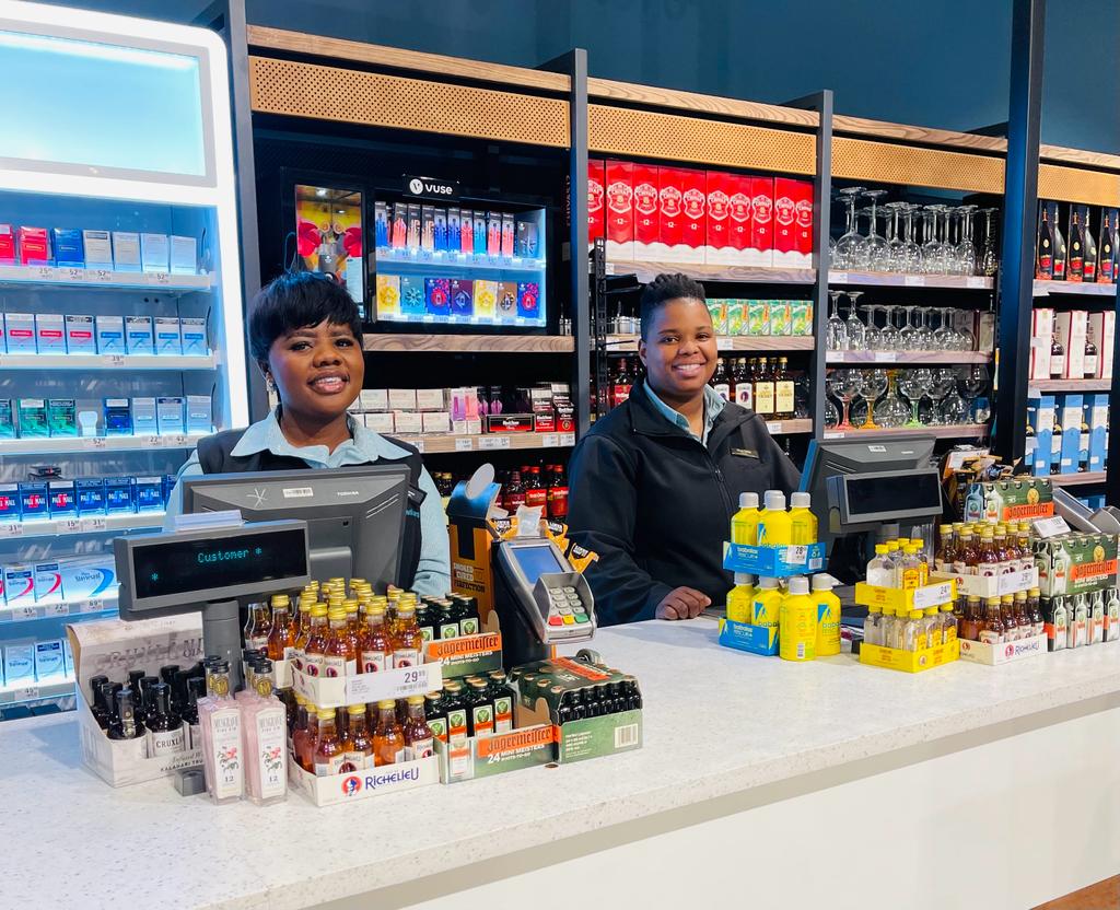 Two smiling checkout clerks stand behind a store counter with drinks and snacks on display shelves behind them.