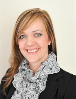 Portrait of a smiling woman with blonde hair, wearing a black blazer and light gray patterned scarf against a pale background.