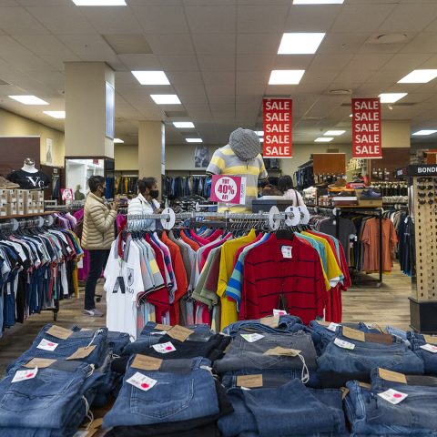 Shoppers browse racks of casual clothing in a busy department store during a sale; foreground shows stacked jeans and colorful shirts on display.