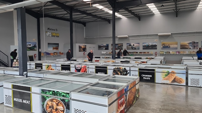 Indoor market or showroom with long rows of freezer units displaying seafood and meat; signs read Mussel Meat, Crumbled Fillets, and other products; customers browse.