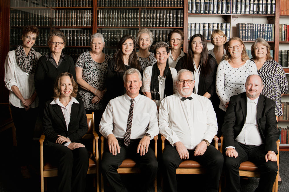 Group of fifteen professionals in a library, seated and standing, smiling at the camera with tall bookshelves behind them.