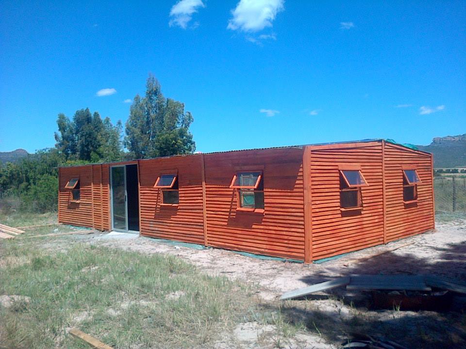 Long, orange-brown wooden modular building under construction in a rural field with small angled windows.