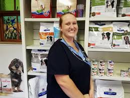 Smiling pet store staff member in navy scrubs standing beside shelves of dog food and supplies.