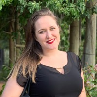 Smiling woman with long light brown hair and red lipstick, wearing a black top, outdoors in a garden.