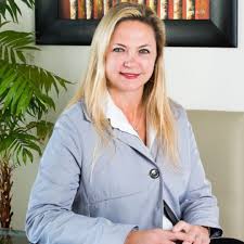Professional woman with blonde hair wearing a light blue blazer, seated in an office and smiling at the camera.