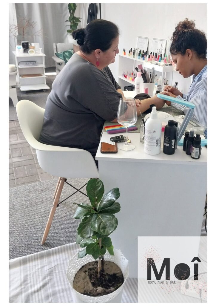 Woman receiving a manicure at a bright nail salon station, with nail tech filing her nails and rows of nail polish bottles in the background.