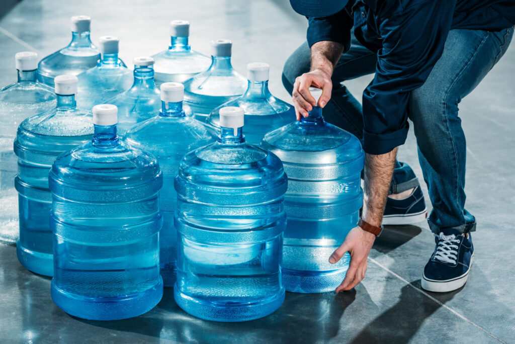 Person crouching among stacked blue 5-gallon water jugs, adjusting the cap on one jug in a storage area