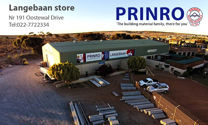 Aerial view of a large green warehouse with PRINRO and LANGEBAAN store banners, building-material yard with stacked pipes and cars nearby, contact details visible on the left.