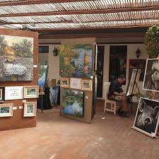 Outdoor art market stall with large map paintings on wooden panels and visitors browsing under a woven roof.