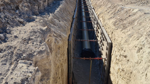 Long construction trench with a large pipe laid along the bottom and steel formwork panels on the right, surrounded by dirt walls.