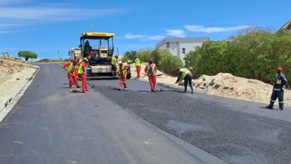 Road construction: workers in safety vests lay asphalt with a paver on a residential street, with equipment nearby.
