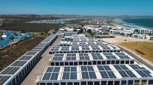 Row after row of solar panels on flat roofs in a coastal industrial area near the sea and beach area.