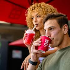 Two friends sharing bright red drinks with straws, standing close outdoors.
