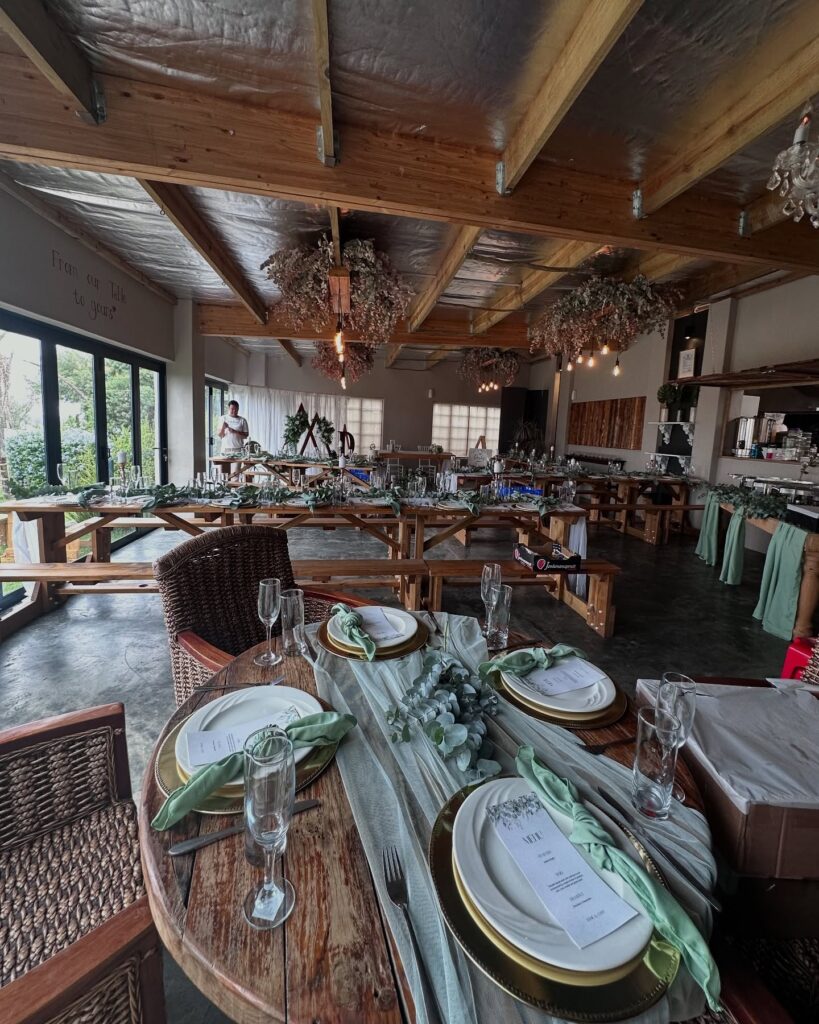 Rustic event hall set for a wedding reception with long wooden tables, greenery runners, and hanging dried flower arrangements under a wooden ceiling.