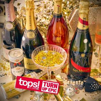 Celebration scene with multiple champagne bottles, a glass dish of gold confetti, and a 'tops! Turn Up Epic' sticker on a festive table.