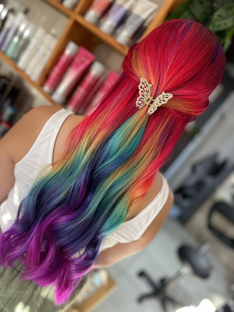 A new layer hair beauty studio – – Iiback view of a person with long, rainbow-colored hair tied with a jeweled butterfly clip in the center, wearing a white tank top in a hair salon setting