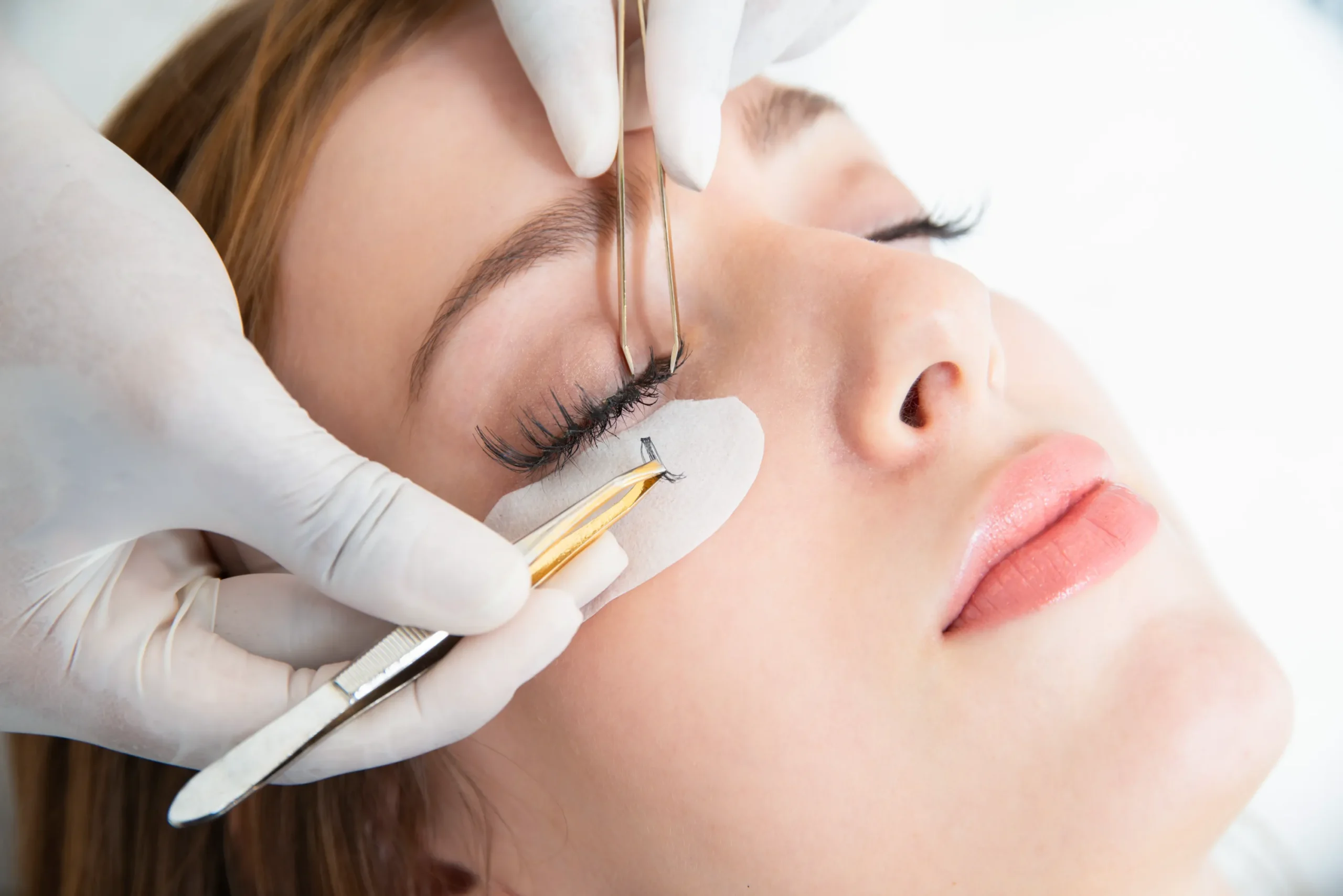 Iiclose-up of a cosmetic procedure near the eye, gloved hands use tweezers near the eyebrow and eyelid with a protective pad under the eye.