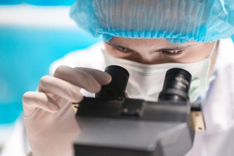 Iiresearcher in a blue surgical cap and mask looks through a microscope in a lab, inspecting slides.