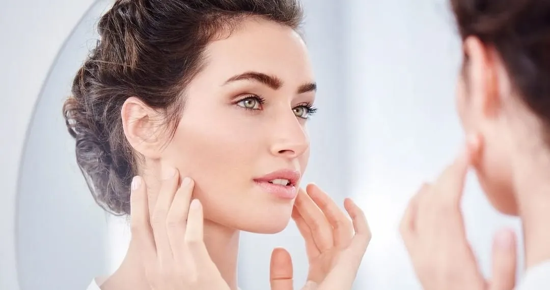 Iiwoman gazing at her reflection in a mirror while touching her face with both hands, as if inspecting her skin for skincare.
