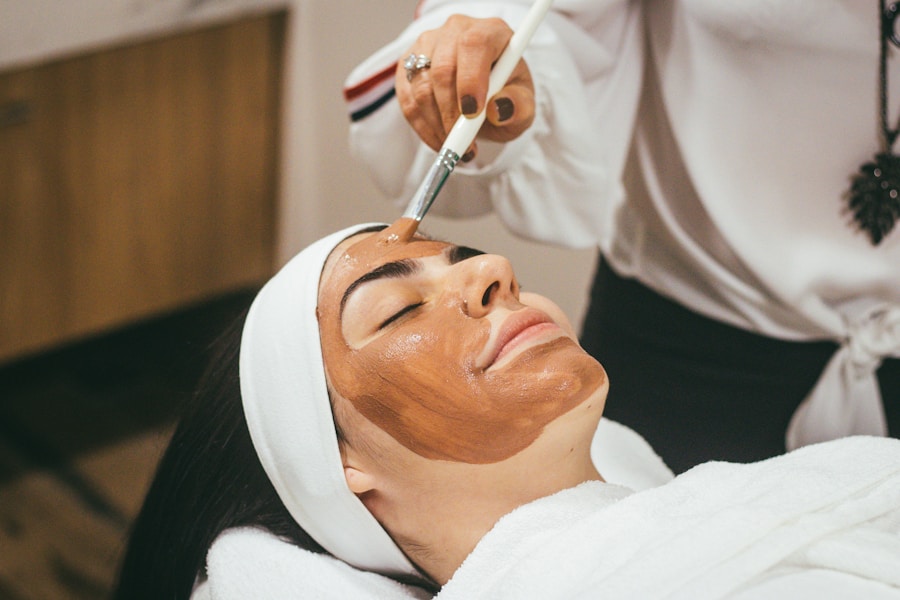 Wellness-skincare-1 jpg – – Iiwoman receiving a brown facial mask application with a brush during a spa treatment, eyes closed and head wrapped in a towel cool and relaxed setting.