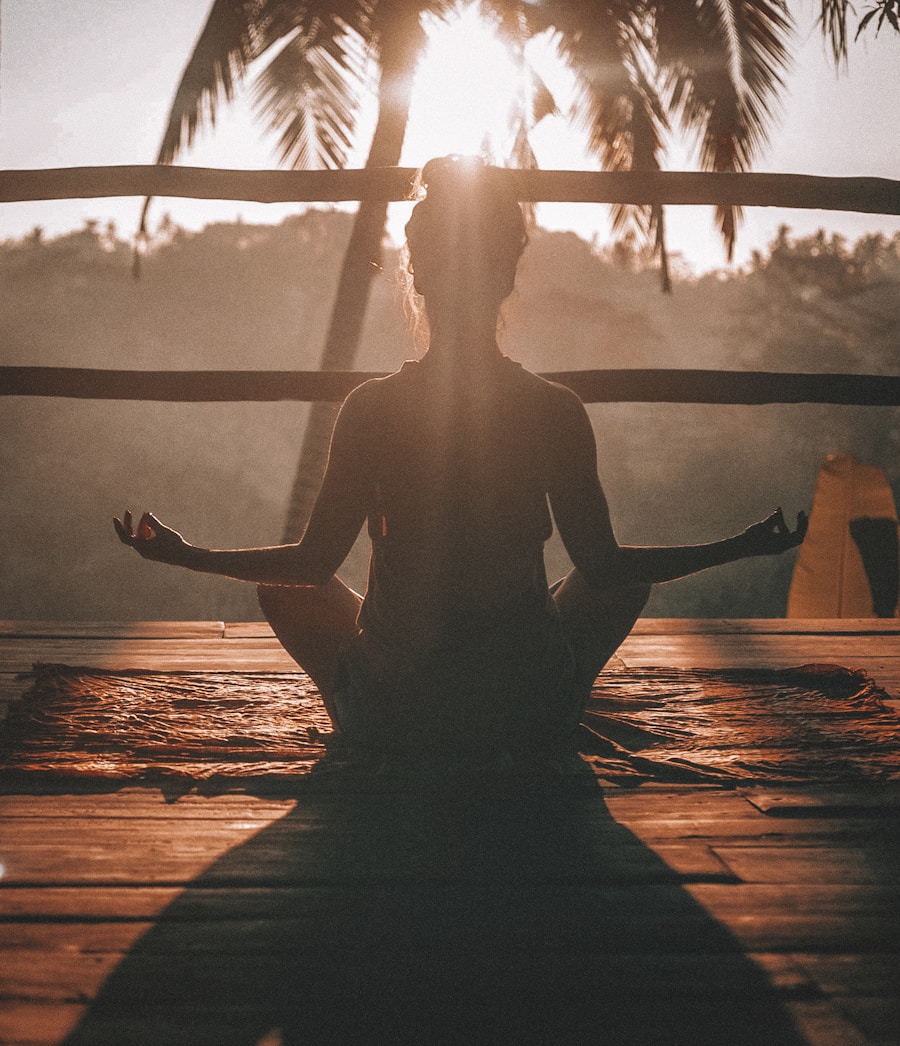 Wellness-yoga-1 jpg – – Iisilhouetted person meditating in a cross‑legged pose on a wooden deck at sunset, with palm trees in the background. (informative)