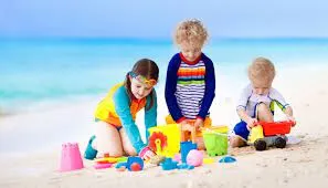 Iithree children playing with colorful sand toys on a sunny beach, with turquoise sea in the background.
