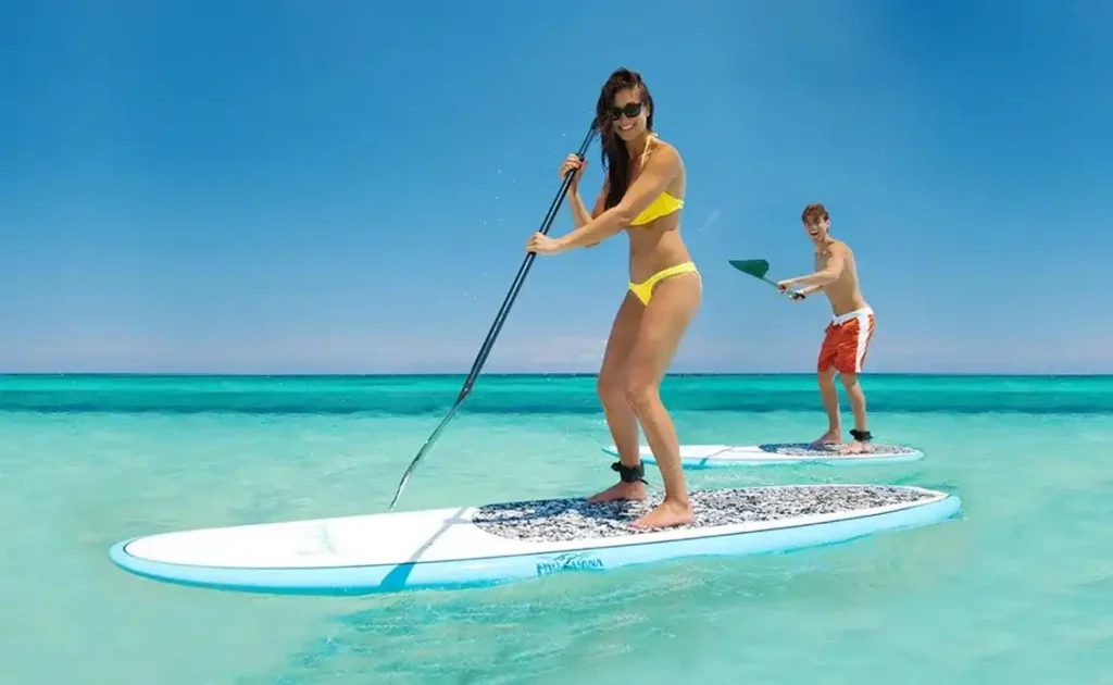 Iitwo people stand-up paddleboarding on turquoise water; woman in yellow bikini in the foreground and a man in orange shorts in the background under a clear blue sky.