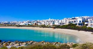 Iicoastal resort town with white buildings along a curved sandy beach and turquoise water under a bright blue sky.