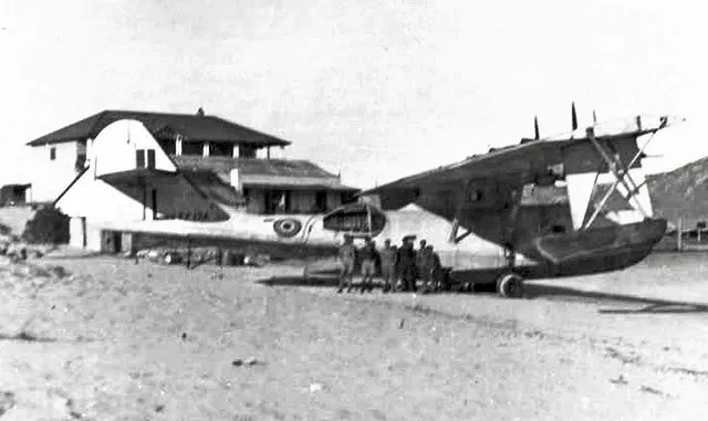Iigroup of people standing beside two vintage propeller aircraft on a dusty airfield with a hangar in the background.